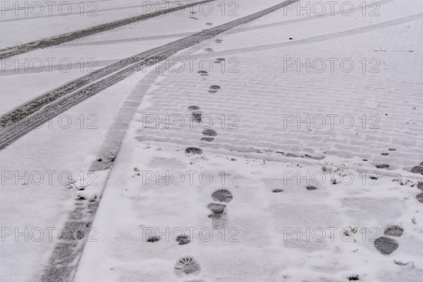Fresh snow covers a cobblestone street with footprints and tire tracks crossing, creating a winter