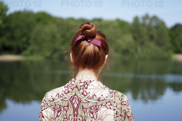 back view of unrecognizable young woman with floral dress and red hair bun looking at lake in