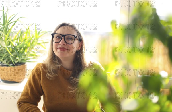 Portrait of smiling middle-aged woman wearing glasses sitting in room with green potted