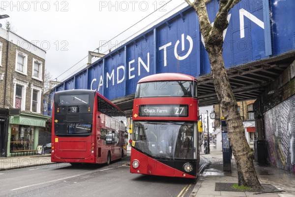 Red double decker buses driving on a london street, passing under a bridge with the camden town