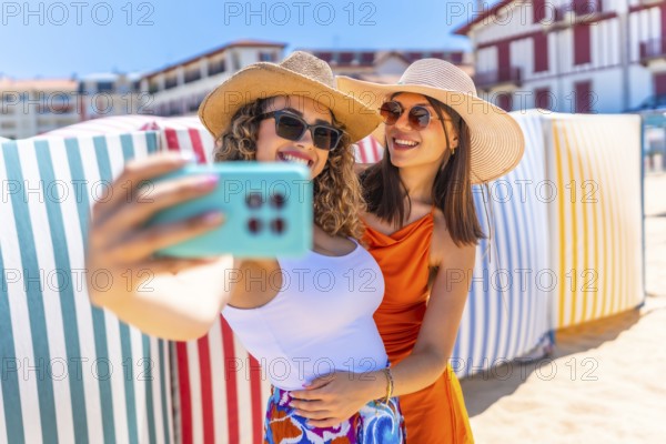 Two smiling women with sun hats and sunglasses taking a selfie on a sunny beach with colorful