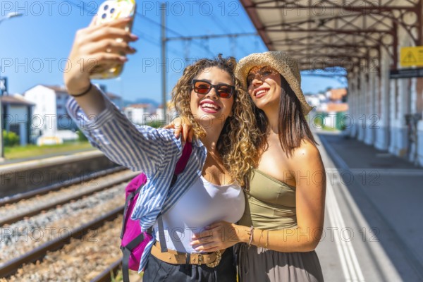 Two happy female tourists taking a selfie with smartphone at a train station, enjoying their summer