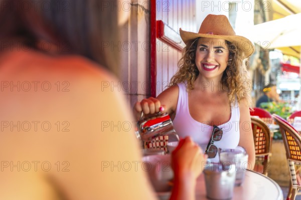 Smiling tourist wearing straw hat pouring milk into coffee, having breakfast with friend at outdoor
