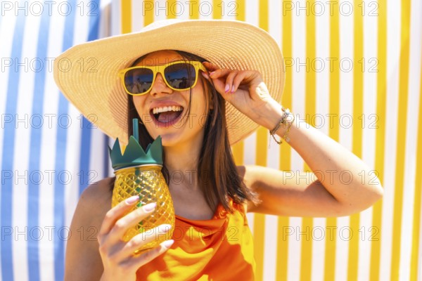 Young woman with sunglasses and a straw hat, enjoying a tropical pineapple juice on a colorful