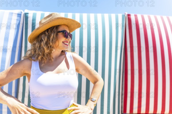Young woman with curly hair, sunglasses, and straw hat posing with hands on hips in front of