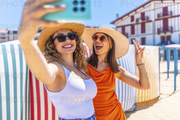 Two cheerful women wearing sun hats and sunglasses, capturing a fun selfie on a sunny beach,