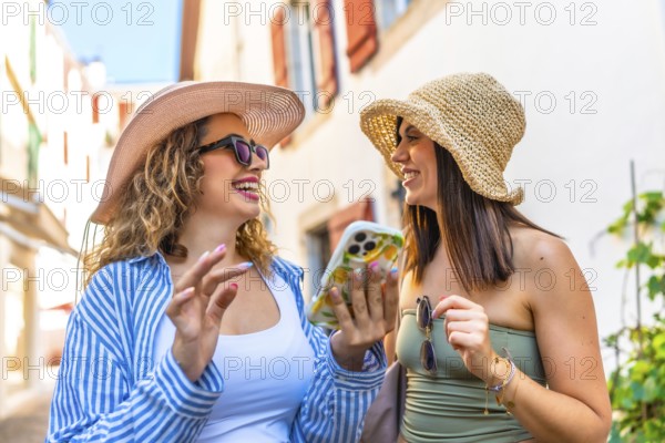 Two happy women wearing summer hats and sunglasses are using a smartphone and laughing while