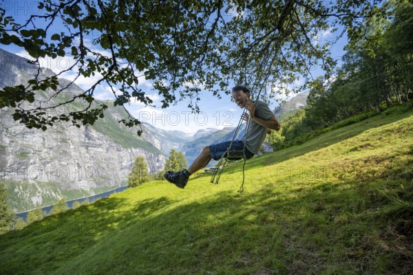 Young man swinging, Blomberg Gård, above the Geirangerfjord, view of idyllic fjord landscape, near