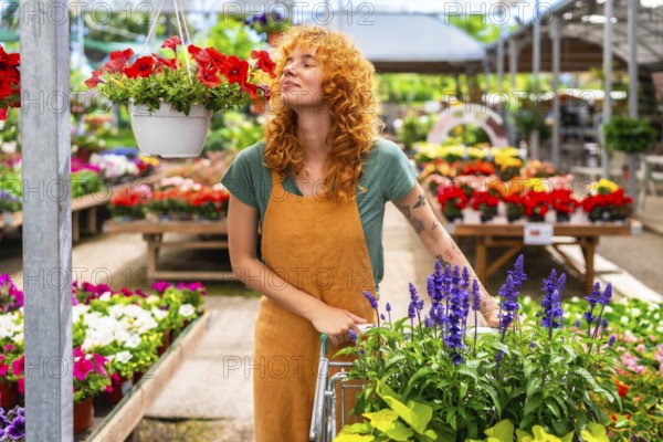 Redhead woman gardener smelling red petunias while pushing a cart in a colorful greenhouse full of