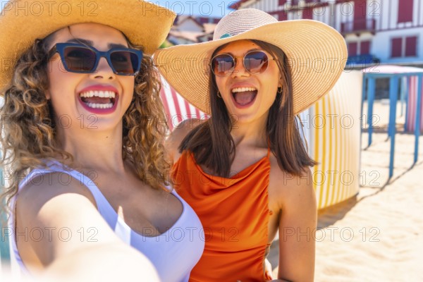 Two cheerful women wearing sunglasses and summer hats are taking a selfie on a sunny beach with