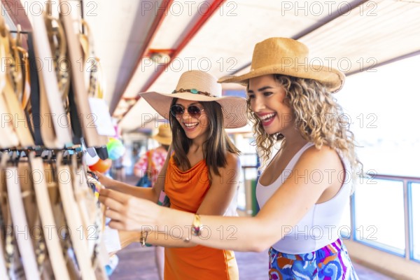 Two smiling women wearing summer hats and sunglasses are choosing souvenirs at a seaside shop while