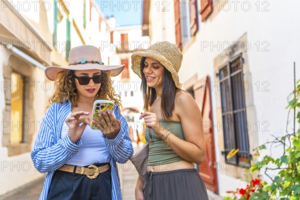 Two female tourists wearing summer hats, using smartphones while walking through charming old town