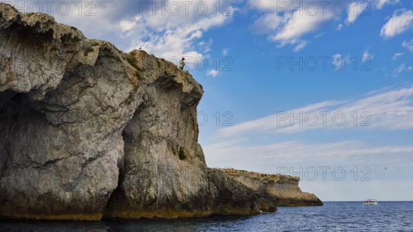 Person fishing from a rocky cliff under a cloudy sky, boat trip around the island of Ortigia, Isola