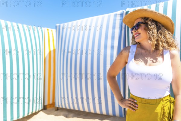 Young woman with curly hair, sunglasses and straw hat posing on beach with colorful striped cabanas