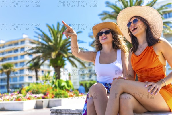 Two happy female tourists wearing summer dresses and straw hats pointing at something while