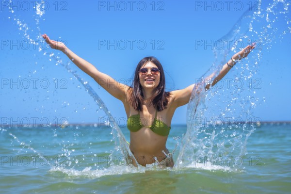 Young woman wearing bikini and sunglasses splashing water with open arms in the sea, enjoying