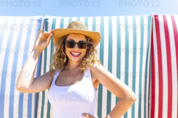 Cheerful young woman with curly hair, wearing sunglasses and a straw hat, smiles while posing
