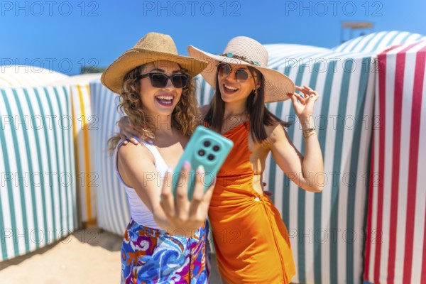 Two happy women wearing summer hats and sunglasses are taking a selfie with a smartphone on a sunny