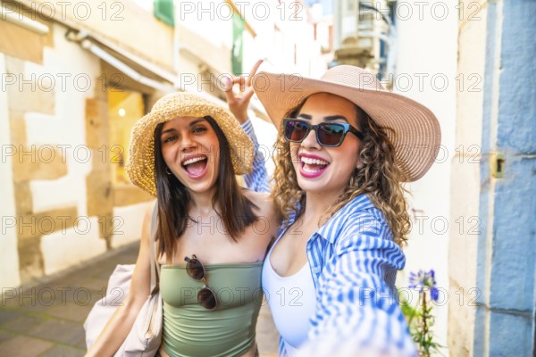 Two cheerful female tourists wearing summer hats and sunglasses are taking a selfie with a cheerful