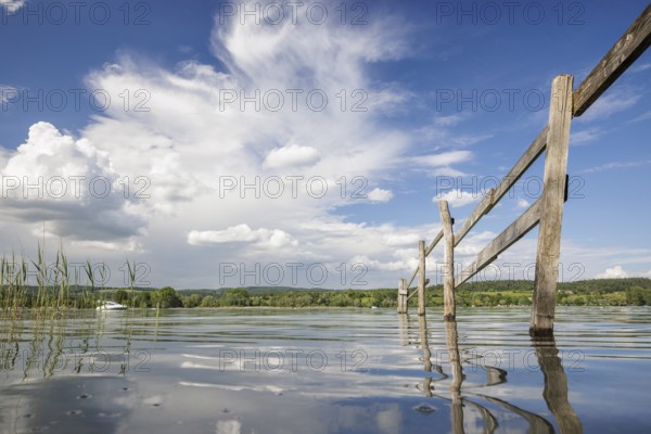A wooden fence stretches into the calm waters of a lake, surrounded by a wide sky and white clouds,