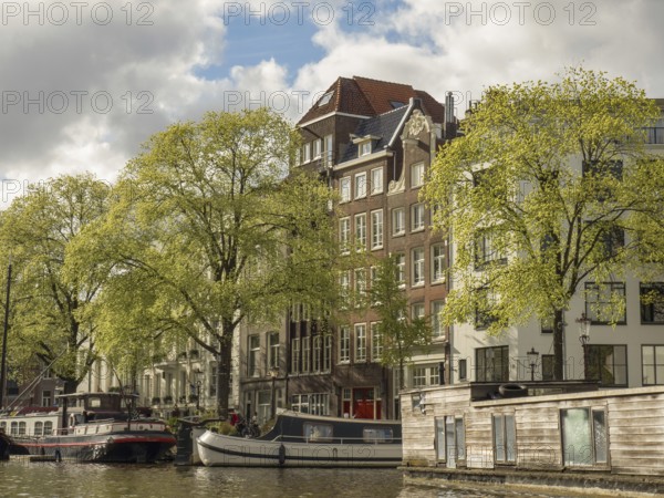 Building fronts along a canal with boats in the water, surrounded by green trees and a cloudy sky,