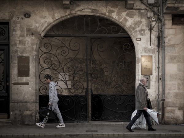 Individuals walk past an arched entranceway with ornate iron gates in central Bordeaux, France,