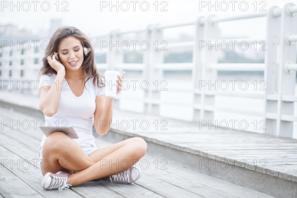 Happy young woman with music headphones and a cup of coffee to go, surfing the internet on a tablet