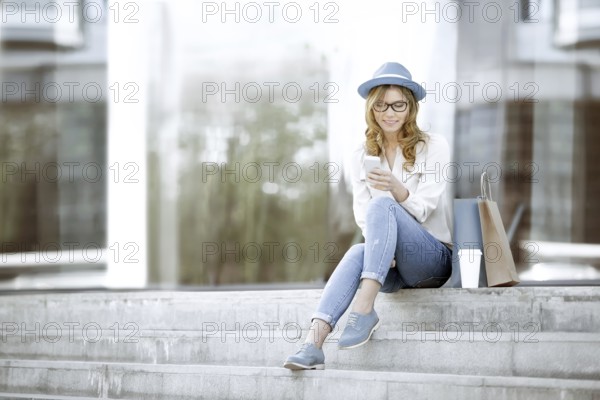 Cheerful young woman with a disposable coffee cup and shopping bags, sitting on the stairs and