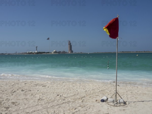 A red warning sign stands on the beach with turquoise water and a blue sky in the background,