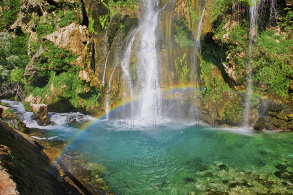 Waterfall with rainbow, surrounded by rocks and green foliage under a clear sky, Topoljski Buk