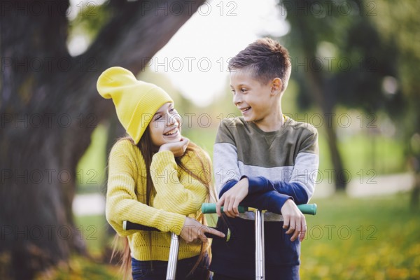 Two children caucasian boy and girl twins posing with scooters in autumn park. Happy kids riding