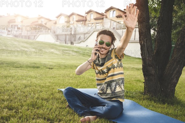 Young stylish handsome man is sitting on grass, resting and talking on the phone. waves his hand