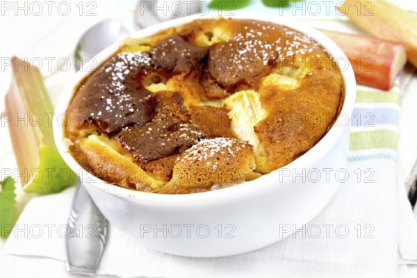 Pie klafouti with rhubarb in the bowl, mint and rhubarb stalks on a napkin against light wooden