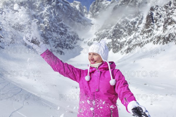 Young woman in pink ski jacket and white gloves smiling and throwing snow in the air with mountain