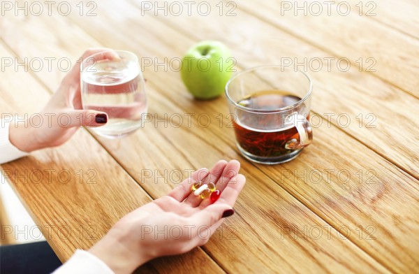 High angle of anonymous female with pills and cup of water witting at table with tea and apple