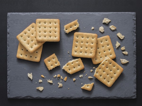 Square crackers with pieces and crumbs on slate gray background. Dry salt cracker cookies with