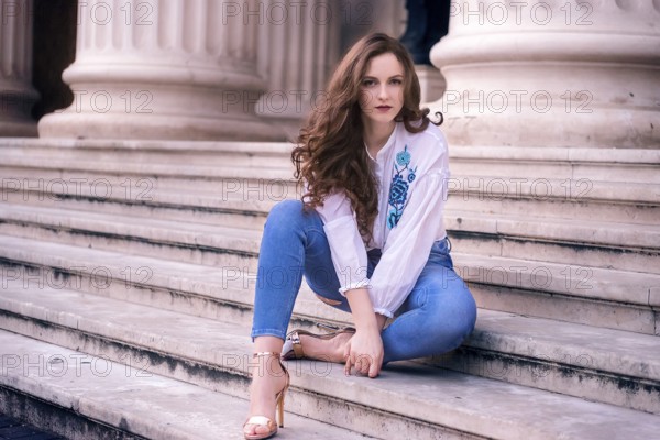 Young girl with brown hair and blue eyes, sits on old marble steps, dressed modern and looking at