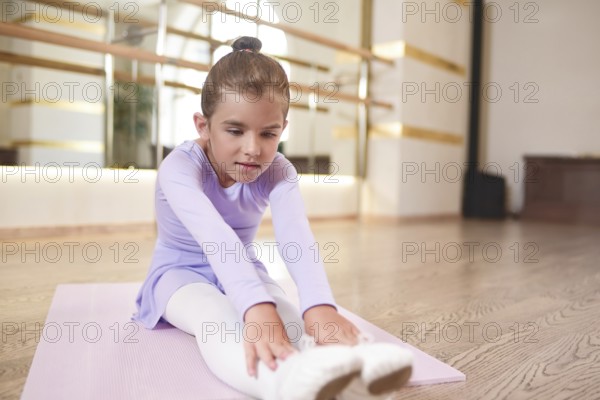Adorable photo of little ballerina in a lilac dress and white tights stretching exercises in a hall
