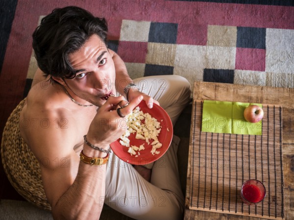 From above shot of happy muscular shirtless man sitting on floor having breakfast eating cereal,