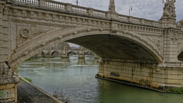 A grand stone bridge with intricate carvings spans over a calm river beneath a cloudy sky, Vittorio