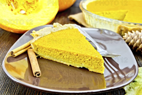 A piece of pumpkin pie in a brown plate with a fork and cinnamon, pumpkin, cones and maple leaf on