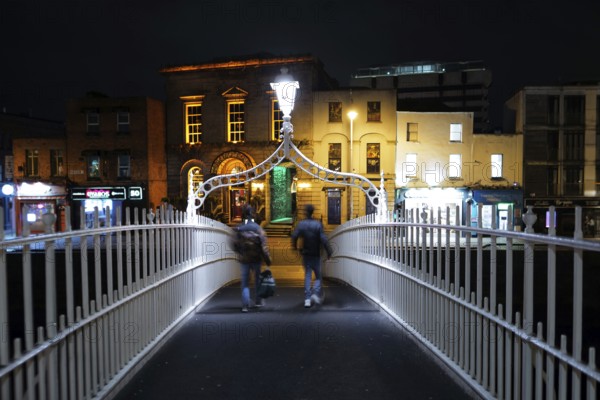 A view of two adults crossing the Ha'penny Bridge on the River Liffey near Temple Bar at night-time