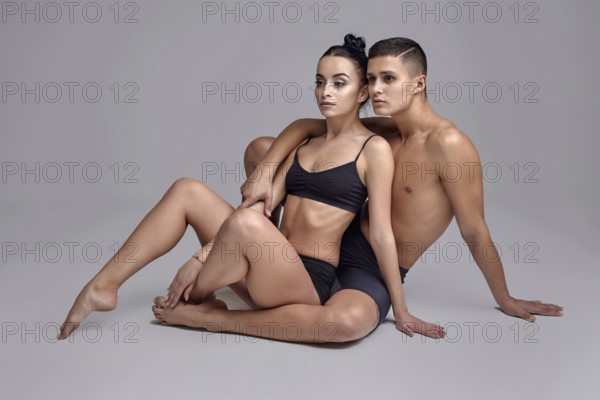 A pair of a graceful athletic ballet dancers in black suits are posing over a gray studio