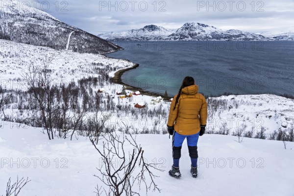 A woman in winter clothing stands above the small bay of Skjellvika on the island of Kvaloya,