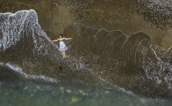 Woman with swimsuit and white dress resting on a sandy beach with breaking waves on the shore.