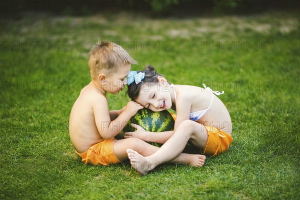 Two children, Caucasian brother and sister, sitting on green grass in backyard of house and hugging