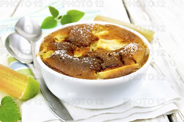 Pie clafouti with rhubarb in the bowl, mint and rhubarb stems on a napkin on wooden plank