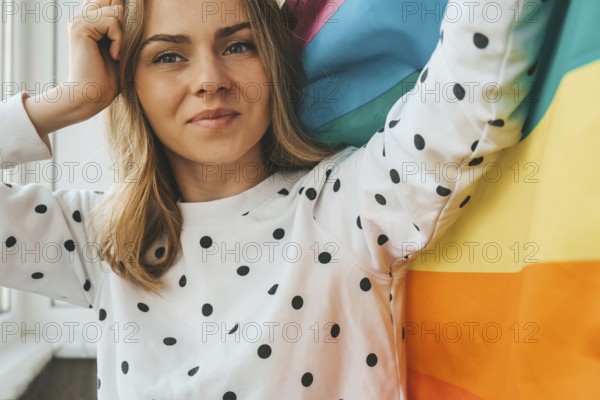 Young blonde millennial hippie woman with rainbow LGBTQ flag at home balcony. Peace and freedom the