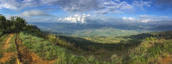 Panorama over the cliffs into a green valley and mountains in sun and shadow, blue sky and white