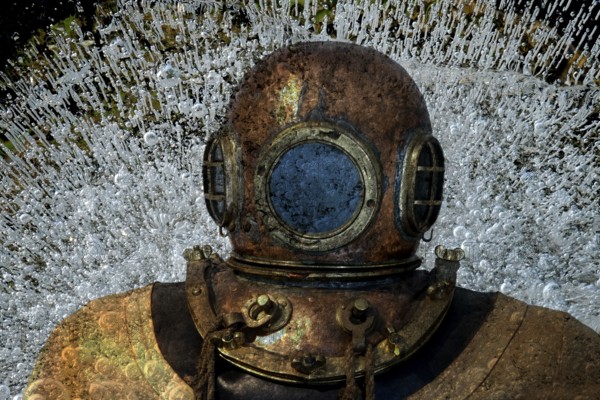 Metal diving helmet with round viewing windows amidst swirling air bubbles. Landscape format.